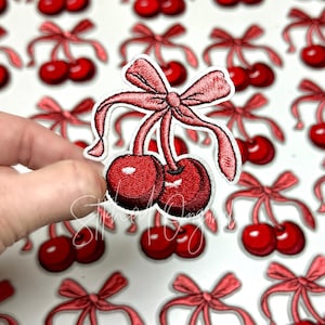 May include: A set of embroidered iron-on patches featuring red cherries with pink bows. The cherries are a classic design and would be perfect for adding a touch of whimsy to any project.