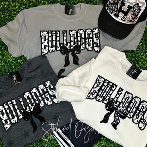 May include: A collection of apparel featuring the word "BULLDOGS" in a distressed, white font with a black bow accent. Items include a gray t-shirt, a dark gray star-patterned t-shirt, and a white sweatshirt. A black and white baseball cap is also included.