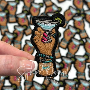 May include: A brown hand holding a blue and white margarita glass with a lime wedge. The hand is wearing a turquoise and silver bracelet. The background is a white surface with many of the same patches.