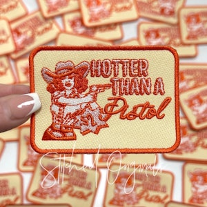 May include: A beige embroidered patch with an orange border. The patch features a cartoon woman in a cowboy hat holding a gun and the text "Hotter Than A Pistol".