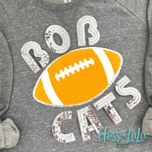 May include: Gray sweatshirt with a silver sequin design that says "BOB CATS" with a football in the middle. The football is orange with white stitching and a white outline. The sweatshirt also has the text "Hoss + Lulu Stitched" at the bottom.