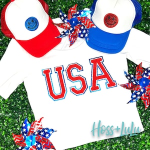 May include: White t-shirt with red, white, and blue glitter "USA" lettering. The shirt is surrounded by red, white, and blue pinwheels and two red, white, and blue trucker hats with smiley faces.