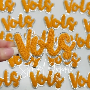 May include: Close-up of several orange felt patches with the word "Vols" embroidered in white. The patches have a silver glitter trim.