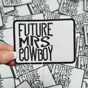 May include: A white rectangular patch with black embroidered text that reads "FUTURE MRS. COWBOY." The patch has a black border and is held by a hand. The background shows other similar patches.