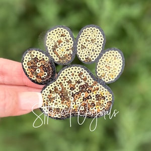 May include: A gold glitter paw print iron-on patch. The paw print is made of gold sequins on a black felt backing.
