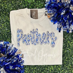 May include: A cream-colored t-shirt with the word "Panthers" in blue and silver sequins. The text "Stitched Originals" is written in cursive below the word "Panthers". Blue and silver pom-poms are on the sides.