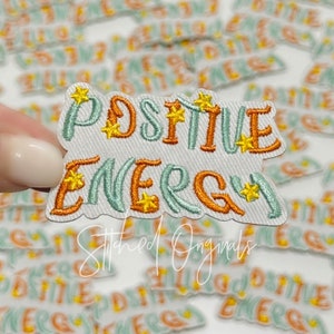 May include: A white fabric patch with the words "POSITIVE ENERGY" embroidered in orange and light blue with yellow star accents. The patch is surrounded by other similar patches with the same design.