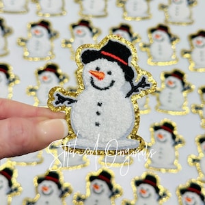 May include: A white snowman with a black top hat and red trim, embroidered with a carrot nose and black eyes. The snowman is surrounded by a gold glitter outline and is on a white background.