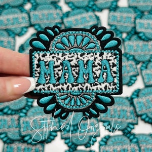 May include: A turquoise and white embroidered patch with the word "MAMA" in a bold font. The patch has a black border and a decorative design with a silver glitter accent.