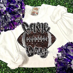 May include: White sweatshirt with a brown and silver sequined football and the text "Game Day" in black sequins.