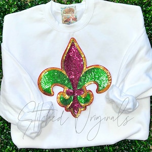 May include: White sweatshirt with a large sequined fleur-de-lis in green, pink, and gold. The sweatshirt has the text "Stitched Originals" printed on the bottom.