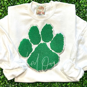 May include: White sweatshirt with a green paw print design. The paw print is outlined in silver glitter.