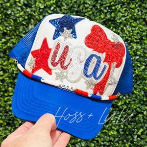 May include: A white trucker hat with a red, white and blue "USA" design. The hat has a blue mesh back and a blue brim. The hat is decorated with sequins, stars and a red bow.