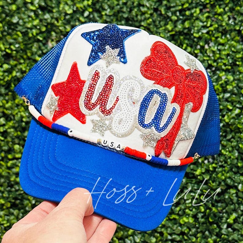 May include: A white trucker hat with a red, white and blue "USA" design. The hat has a blue mesh back and a blue brim. The hat is decorated with sequins, stars and a red bow.