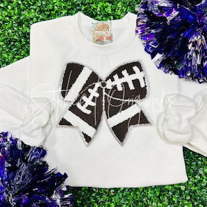 May include: White sweatshirt with a brown and white football-themed bow. The bow is made of a fuzzy material and has a silver glitter outline.