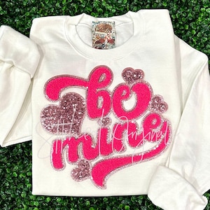 May include: White sweatshirt with a pink and rose gold glitter design that says "Be Mine".
