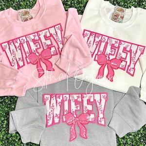 May include: Three sweatshirts with the word "WIFEY" in white with a pink floral pattern and a pink bow. The sweatshirts are in pink, white, and gray.