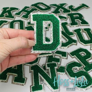 May include: Green felt letter patches with a white outline and silver glitter trim. The letters are D, A, N, and R. The letters are arranged in a pile. The letters are for sale on Etsy.