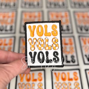 May include: Embroidered patch with the word "VOLS" in orange, orange and white checkered, and black lettering. The patch has a black border and is on a white background. The patch is held in a hand.