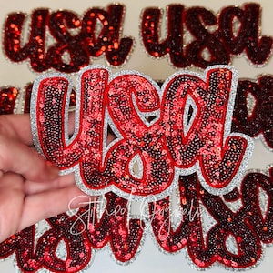 May include: Red sequined "USA" patch with a silver glitter border. The letters are in a bold, cursive font. Several patches are visible, with one held up in front of others.