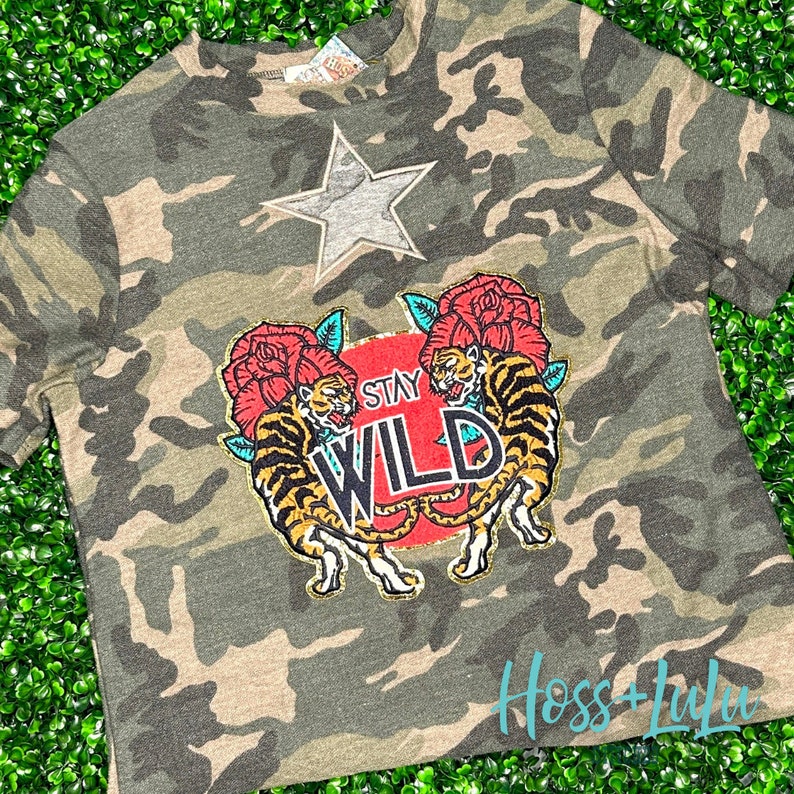 May include: A camouflage print t-shirt with a patch featuring two tigers, roses, and the text "Stay Wild".