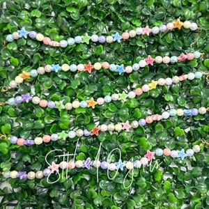 May include: A set of colorful beaded bracelets with star-shaped charms. The bracelets are made with pastel-colored beads and feature a variety of colors, including pink, blue, yellow, and green. The star charms are also colorful and add a playful touch to the bracelets.