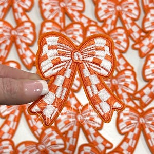 May include: An orange and white checkered bow patch with an embroidered design. The bow is held in front of a background of similar bows. The bow has a detailed, textured appearance.