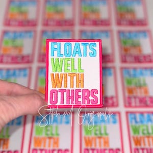 May include: Embroidered patch with the text "FLOATS WELL WITH OTHERS" in blue, green, orange, and pink. The patch has a white background and a pink border. The words are stacked vertically. The patch is held in front of a background of similar patches.