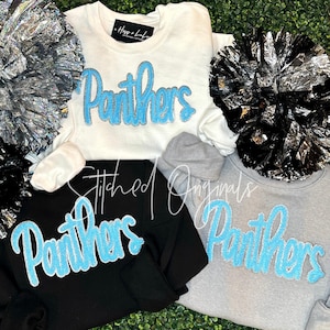 May include: White, black, and gray sweatshirts with the word "Panthers" in light blue glitter lettering. The sweatshirts are surrounded by silver and black pom-poms. The brand name "Hoss + Lulu" is visible on the white sweatshirt.