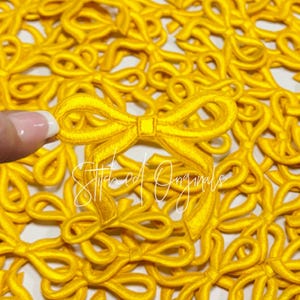 May include: A close-up of a pile of yellow fabric bows. The bows are all the same size and shape, and they are arranged in a random pattern. The bows are made of a soft, silky fabric.