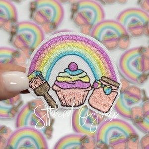 May include: A rainbow embroidered patch with a cupcake, paintbrush, and jar. The cupcake is pink, yellow, and green. The paintbrush is brown and gold. The jar is pink and blue. The rainbow is pink, yellow, orange, green, blue, and purple.