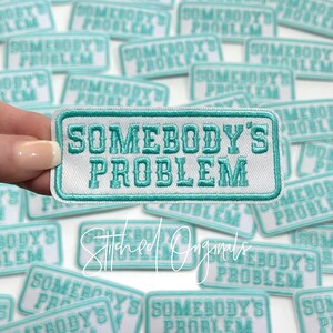 May include: A white patch with teal blue embroidered text that reads "Somebody's Problem". The patch is surrounded by other similar patches.