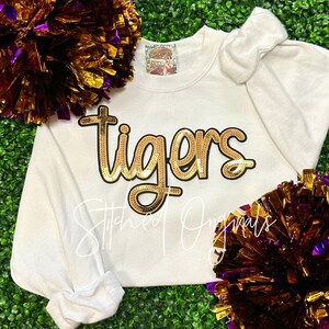 May include: Cream-colored sweatshirt with the word "tigers" in gold sequin lettering. The sweatshirt has long sleeves and a crew neck. The image also includes gold and purple pom-poms, and the text "Stitched Originals".