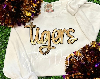 May include: Cream-coloured sweatshirt featuring the word "tigers" in gold sequin lettering. The long-sleeved top has a crew neck. The image also includes gold and purple pom-poms, and the text "Stitched Originals".