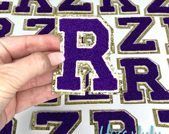 Könnte beinhalten: Lilafarbene Filzbuchstaben-Patches mit goldfarbenem Glitzerbesatz. Der Buchstabe R wird in einer Hand gehalten. Die Patches sind zum Nähen oder Bügeln auf Kleidung oder andere Gegenstände geeignet.