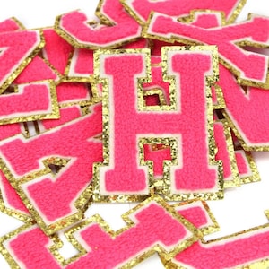 May include: A pile of pink felt letters with gold glitter trim. The letters are in a variety of sizes and styles, and they are all uppercase.
