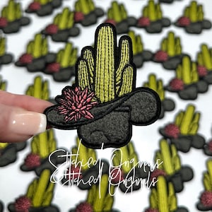 May include: A black felt cowboy hat with a pink flower and a cluster of green cacti embroidered on top. The hat is surrounded by other similar patches.