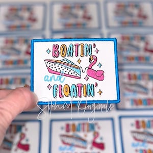 May include: Embroidered patch with a blue border featuring a boat and a flamingo, with the words "BOATIN' and FLOATING" in colorful text. The patch is white with a blue border and is held up by a hand.