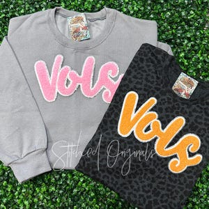 May include: Two gray and black sweatshirts with the word "vols" in pink and orange fuzzy letters. The sweatshirts are on a green background.