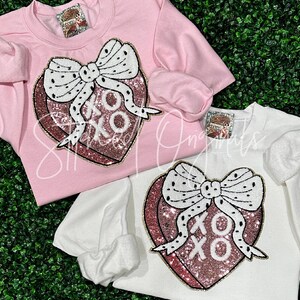 May include: Two pink and white sweatshirts with a pink and gold heart with a black and white bow and the text "XO" on it.