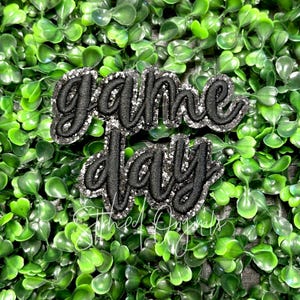 May include: Black and silver glitter iron-on patch with the text "game day" in a cursive font.