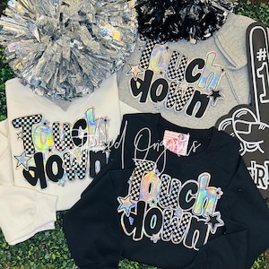 May include: Four sweatshirts in white, gray, and black, each with the word "Touchdown" in a holographic, checkered, and star-studded design. Silver and black pom-poms and a foam finger are also visible.