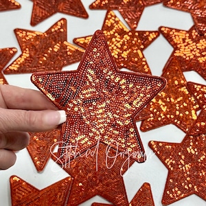 May include: A close-up of a collection of orange star-shaped patches with sequins. The patches are made of a sparkly fabric and have a variety of designs. One patch has the text "What's your story?" written on it.