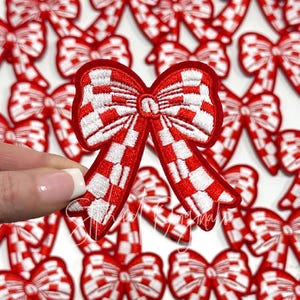 May include: A close-up of a red and white checkered bow, with a hand holding the bow in front of a background of many other bows.
