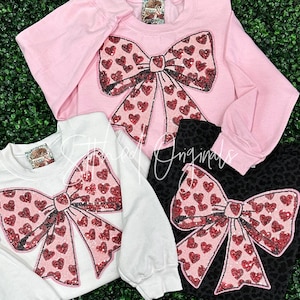 May include: Three sweatshirts with a large red and pink sequined bow with hearts on the front. The sweatshirts are pink, white, and black.