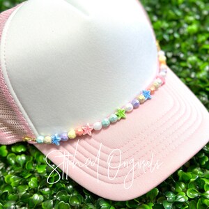 May include: A pink and white trucker hat with a colorful beaded chain featuring pastel-colored beads and star-shaped charms. The hat has a mesh back and a pink brim.