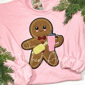 May include: Pink sweatshirt with a gingerbread person holding a pink cup and a yellow rolling pin. The gingerbread person has a red bow tie and a smiling face.