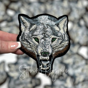May include: A close-up of an embroidered iron-on patch featuring a snarling wolf's head with green eyes. The wolf's fur is white and gray, and the background is black.