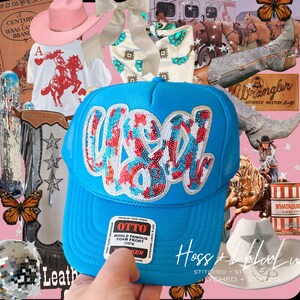 May include: A blue trucker hat with the word "USA" in sequined red, white, and blue letters. The hat is surrounded by other western-themed items, including a pink cowboy hat, a white t-shirt with a horse graphic, and cowboy boots.