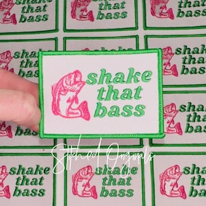 May include: Embroidered patch with the text "shake that bass" in green, featuring a pink fish illustration. The patch has a white background with a green border. The image shows multiple patches.